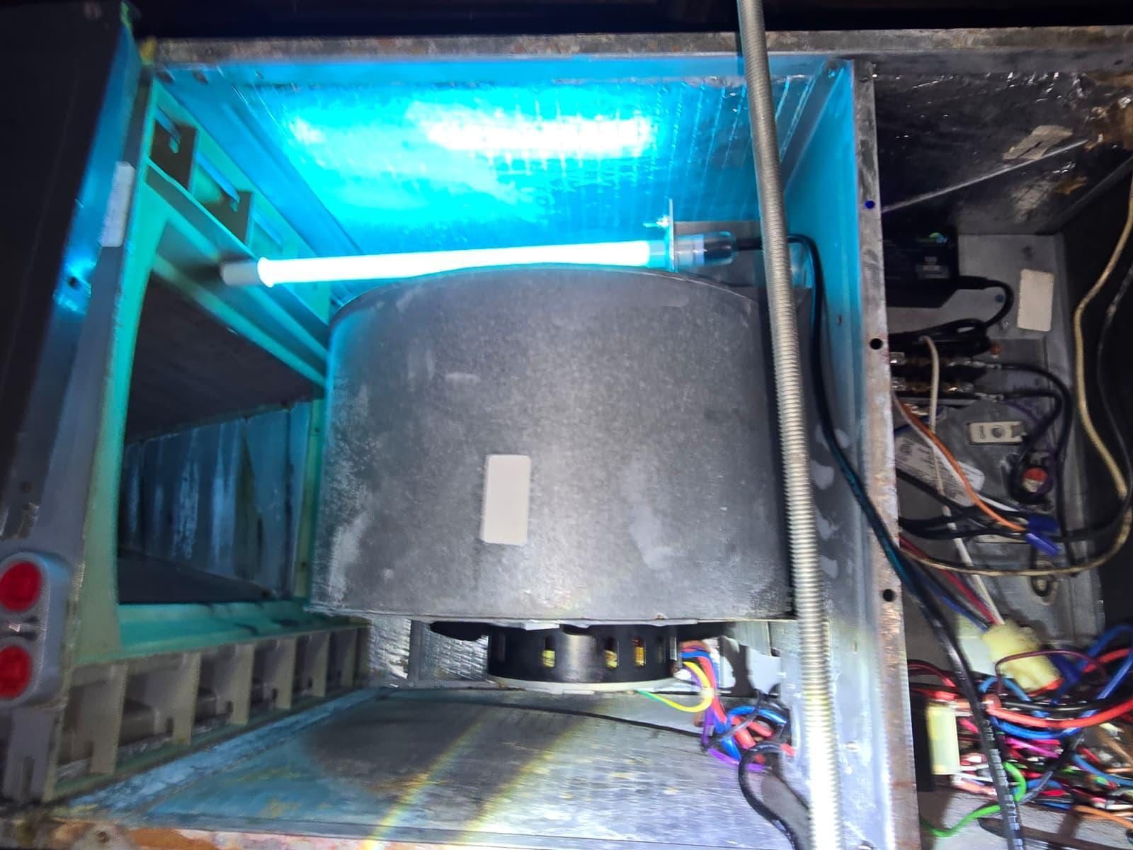 The inside of an air handler cabinet lit by a blue UV lamp, showing a dusty blower housing and the wiring along one side