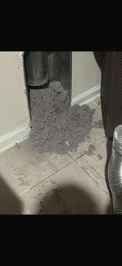 Un montón denso de pelusa gris sacada por la abertura del ducto de la secadora, en el piso junto a un cesto de ropa